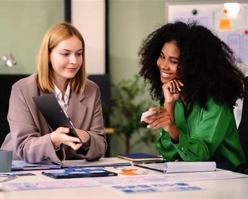 Two women are sitting at a table; one is holding a tablet while the other smiles and points at a map. It's a work environment filled with notes and laptops.