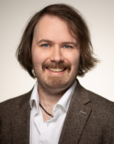 Ein Mann mit einem Bart und lockigem Haar trägt einen grauen Blazer und ein weißes Hemd. Er lächelt freundlich in die Kamera.