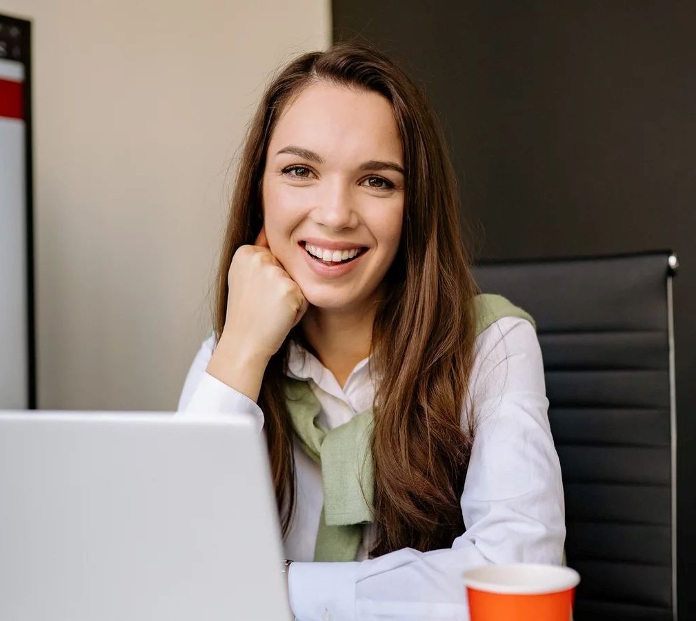 Eine lächelnde Frau sitzt an einem Schreibtisch mit einem Laptop und einer Tasse Kaffee. Im Hintergrund ist ein Bildschirm mit Text zu sehen. Sie trägt ein weißes Hemd und einen grünen Pullover.