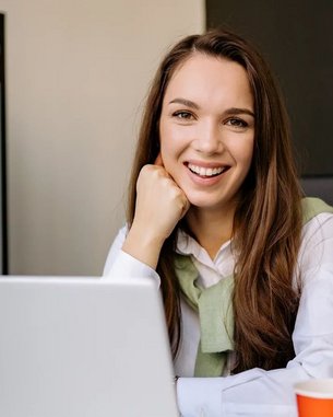 Eine lächelnde Frau sitzt an einem Schreibtisch mit einem Laptop und einer Tasse Kaffee. Im Hintergrund ist ein Bildschirm mit Text zu sehen. Sie trägt ein weißes Hemd und einen grünen Pullover.