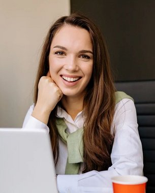 A smiling woman sits at a desk with a laptop and a cup of coffee. In the background, there’s a screen displaying text. She is wearing a white shirt and a green sweater.