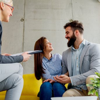 Ein Paar sitzt auf einer gelben Couch und lächelt sich an, während eine Beraterin ihnen gegenübersitzt und Notizen macht.