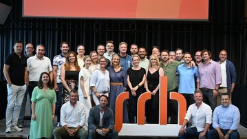Gruppenshot von etwa 40 Personen, die vor einem großen, orangefarbenen Logo stehen. Im Hintergrund steht der Text "Leidenschaft fürs Leben."