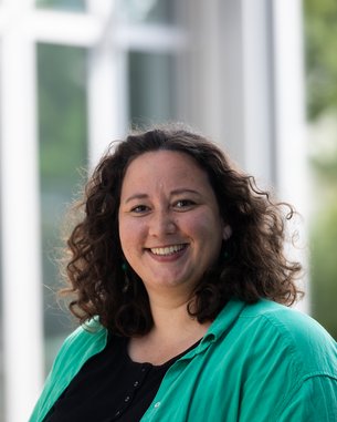 A smiling woman with curly hair is wearing a green shirt over a black top. She is standing in front of a bright background.