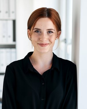 Eine Frau mit braunen Haaren steht an einer Wand in einem modernen Büro, trägt ein schwarzes Hemdkleid und lächelt freundlich.