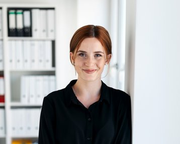 Eine Frau mit braunen Haaren steht an einer Wand in einem modernen Büro, trägt ein schwarzes Hemdkleid und lächelt freundlich.