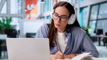 A woman with headphones sits at a desk, focused on her laptop, surrounded by notes and books.