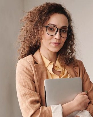 Eine Frau mit lockigem Haar trägt eine Brille und einen beigen Blazer. Sie hält einen Laptop an ihrer Brust und steht in einem modernen Raum. Ihr Gesichtsausdruck ist neutral und professionell.
