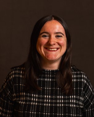 Eine lächelnde Frau mit langen, dunklen Haaren trägt ein kariertes Oberteil. Der Hintergrund ist neutral und unauffällig.