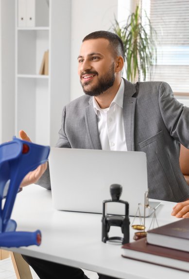 Ein junger Mann hält eine Krücke und spricht mit einem lächelnden Mann in einem Anzug an einem Schreibtisch mit Laptop und Büromaterial.