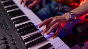 One hand is playing on a keyboard while the other hand is poised to press keys. The person is wearing a watch and sitting on a chair.