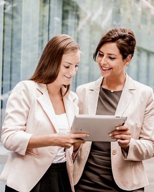 Zwei Frauen in Business-Anzügen stehen draußen und betrachten gemeinsam ein Tablet. Sie lächeln und scheinen sich über den Inhalt des Tablets auszutauschen. Im Hintergrund sind moderne Gebäude und Bäume sichtbar.