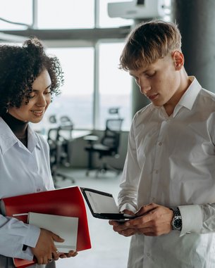 Eine Frau mit lockigem Haar hält einen roten Ordner und ein Blatt Papier, während ein Mann in einem weißen Hemd auf ein Gerät schaut. Beide stehen in einem modernen Büro mit großen Fenstern.
