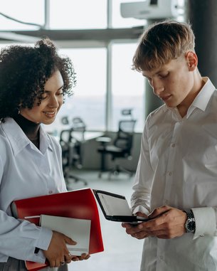 Eine Frau mit lockigem Haar hält einen roten Ordner und ein Blatt Papier, während ein Mann in einem weißen Hemd auf ein Gerät schaut. Beide stehen in einem modernen Büro mit großen Fenstern.