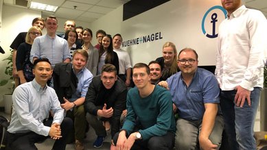 Gruppe von Personen, die in einem Büro stehen, mit einem Schild von Kuehne + Nagel im Hintergrund. Einige Personen posieren für die Kamera.