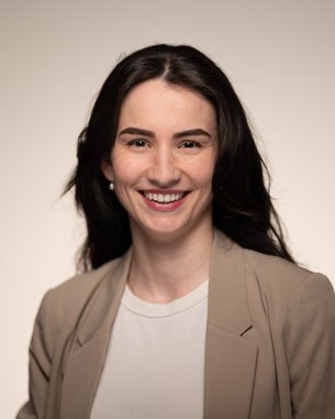 Eine lächelnde Frau mit langen, dunklen Haaren trägt einen beigen Blazer und ein weißes T-Shirt. Der Hintergrund ist neutral und hell.