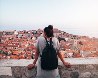 Eine Person mit einem Rucksack steht an einer Mauer und blickt auf eine Stadt mit roten Dächern und dem Meer im Hintergrund. Der Himmel ist klar und blau.
