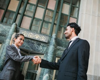 Two individuals in formal attire shake hands in front of a building with large windows and decorative features. The woman is wearing a suit and smiling, while the man is also dressed professionally.