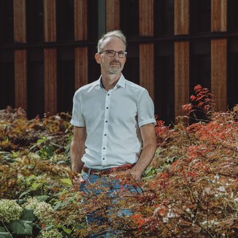 A man stands in a garden filled with colorful foliage and plants. He is wearing a white shirt and gray pants. In the background, wooden panels of a building are visible.