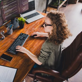 Eine Person mit lockigem Haar sitzt an einem Schreibtisch und arbeitet an mehreren Monitoren, die Programmiercode anzeigen. Auf dem Tisch befinden sich ein Laptop, ein Smartphone und ein Notizblock.