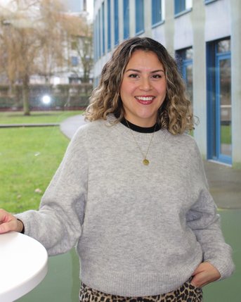 Frau mit lockigem Haar in einem grauen Pullover und Leopardenmuster-Hose steht an einem Tisch in einem modernen Gebäude.