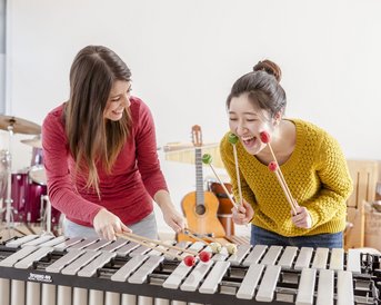 Zwei Frauen spielen gemeinsam auf einem Xylophon und verwenden bunte Schlägel. Im Hintergrund sind Musikinstrumente sichtbar.