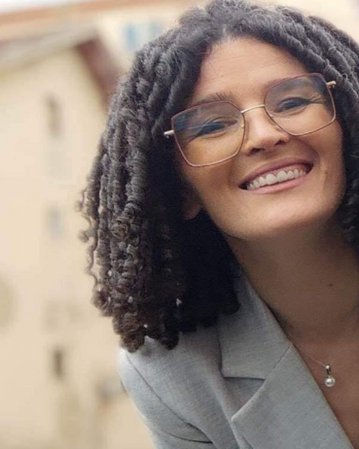 Eine lächelnde Frau mit lockigem Haar und Brille trägt einen grauen Blazer. Der Hintergrund zeigt unscharfe Gebäude.