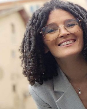 Eine lächelnde Frau mit lockigem Haar und Brille trägt einen grauen Blazer. Der Hintergrund zeigt unscharfe Gebäude.