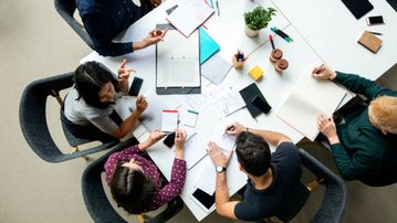 Eine Gruppe von fünf Personen sitzt um einen Tisch, diskutiert und arbeitet an Notizen und Dokumenten. Schreibmaterialien und Smartphones sind sichtbar.