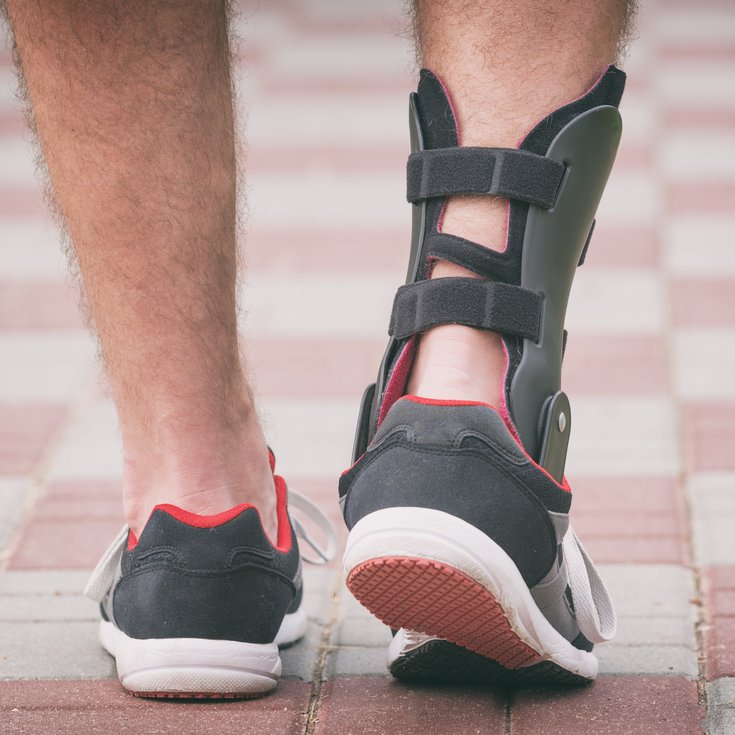 A foot in a sports shoe with a brace on it, walking along a paved path.