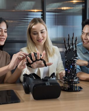 Drei Personen arbeiten an einem Tisch mit Technologie. Eine Frau testet einen Handschuh, während die anderen zuschauen. Auf dem Tisch befinden sich ein VR-Headset und ein Roboterhand-Modell.