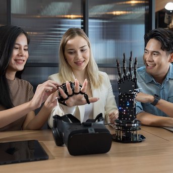 Three people are working at a table with technology. A woman is testing a glove while the others watch. On the table, there is a VR headset and a robotic hand model.