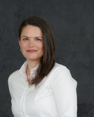 Eine Frau mit langen, braunen Haaren trägt ein weißes Hemd und lächelt leicht vor einem neutralen, grauen Hintergrund.