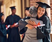 A graduate hugs an older woman while holding her diploma. In the background, other graduates in their caps and gowns can be seen.
