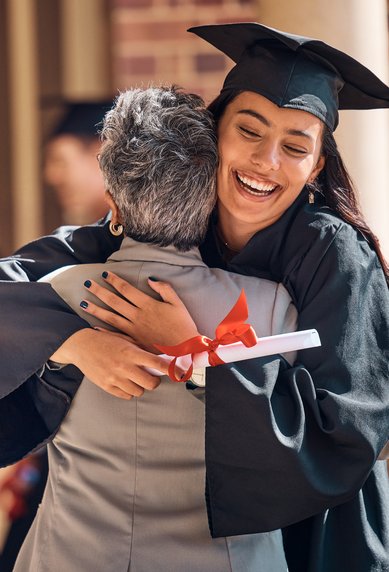 A graduate hugs an older woman while holding her diploma. In the background, other graduates in their caps and gowns can be seen.
