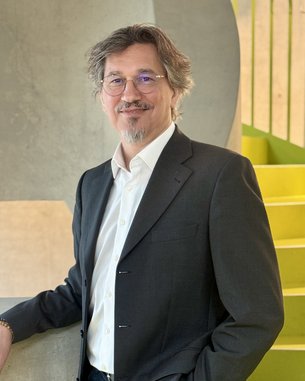 Mann mit Brille und grauem Anzug steht an einer Wand und lächelt. Im Hintergrund sind grüne Treppenstufen sichtbar.