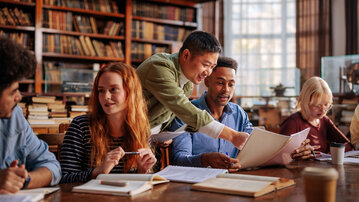 Six people are sitting at a table in a library, discussing documents and notes. Bookshelves are visible in the background.