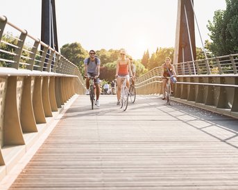 Drei Radfahrer überqueren eine Holzbrücke bei Sonnenuntergang, umgeben von Bäumen und einer ruhigen Umgebung.