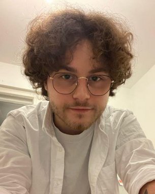 Junger Mann mit lockigem Haar und Brille, trägt ein weißes Hemd, schaut in die Kamera. Helle Innenraumgestaltung im Hintergrund.