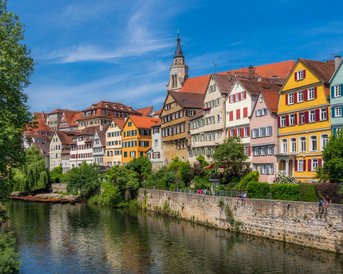 Farbige Fachwerkhäuser entlang eines Flusses, mit Bäumen im Vordergrund und einem Kirchturm im Hintergrund unter blauem Himmel.