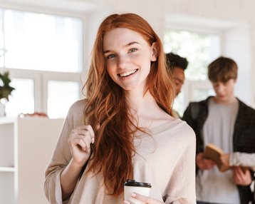 Eine lächelnde Frau mit roten Haaren hält einen Kaffeebecher in einem modernen Raum, während andere Personen im Hintergrund interagieren.