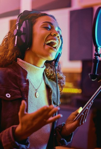 A singer is wearing headphones and standing in front of a microphone in the recording studio, singing while holding a tablet in her hand.