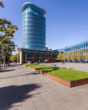 Moderne Architektur mit einem hohen, gläsernen Gebäude und einer gepflasterten Fläche, umgeben von Bäumen und Rasenflächen.