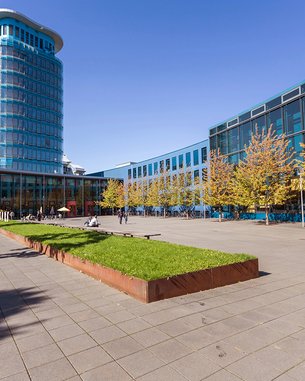 Modern architecture featuring a tall glass building and a paved area, surrounded by trees and grassy lawns.