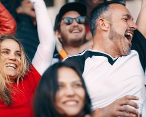 Menschen jubeln und feiern in einem Stadion, gekleidet in Fanbekleidung, während sie ihre Freude über ein Sportereignis ausdrücken.