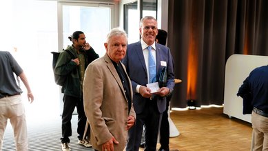 Zwei Männer stehen in einem modernen Raum. Der Mann links trägt einen beigen Blazer, der Mann rechts hält ein Dokument und trägt einen Anzug. Im Hintergrund sind weitere Personen und eine große Fensterfront sichtbar.