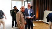 Zwei Männer stehen in einem modernen Raum. Der Mann links trägt einen beigen Blazer, der Mann rechts hält ein Dokument und trägt einen Anzug. Im Hintergrund sind weitere Personen und eine große Fensterfront sichtbar.