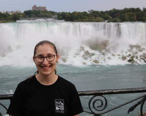 Eine Person mit Brille lächelt vor den Niagarafällen, die im Hintergrund mit Wasserrauschen zu sehen sind.