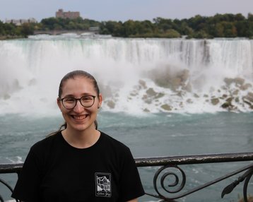Eine Person mit Brille lächelt vor den Niagarafällen, die im Hintergrund mit Wasserrauschen zu sehen sind.