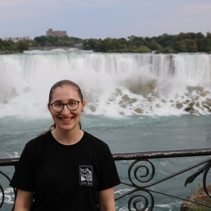 Eine Person mit Brille lächelt vor den Niagarafällen, die im Hintergrund mit Wasserrauschen zu sehen sind.